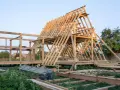 Drewniana konstrukcja domu w budowie. Widoczny szkielet dachu i ścian, schody. Fundament punktowy pod domek drewniany jest już gotowy.