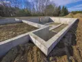 Fundamenty pod dom 120m2 w budowie. Betonowe ściany fundamentowe tworzą zarys przyszłego domu na tle drzew i błękitnego nieba.