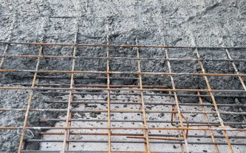 Siatka zbrojeniowa czeka na beton na fundament. Powierzchnia jest szara i chropowata, z widocznymi prętami.