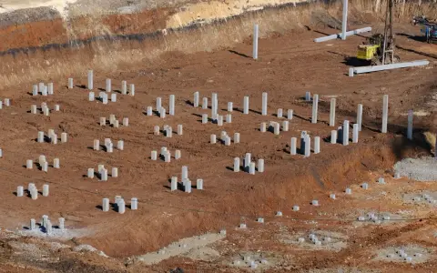 Plac budowy z licznymi betonowymi słupami, tworzącymi fundament pośredni. W tle maszyna budowlana.