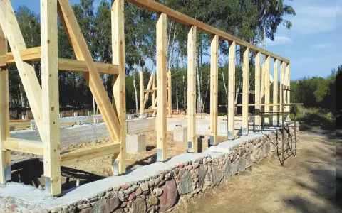 Drewniana konstrukcja domu powstaje na solidnym fundamencie z kamienia. Widać betonowe słupy i kamienny murek.