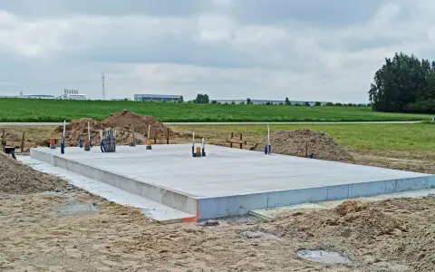 Solidna wylewka pod domek letniskowy gotowa do dalszych prac. Wokół plac budowy z piaskiem i zielone pola w tle.